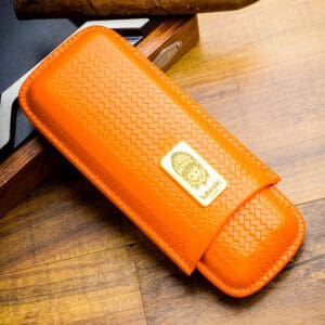 Orange woven leather case with a gold badge on the front, resting on a wood surface. It appears to be a compact accessory or sheath with stitched edges.