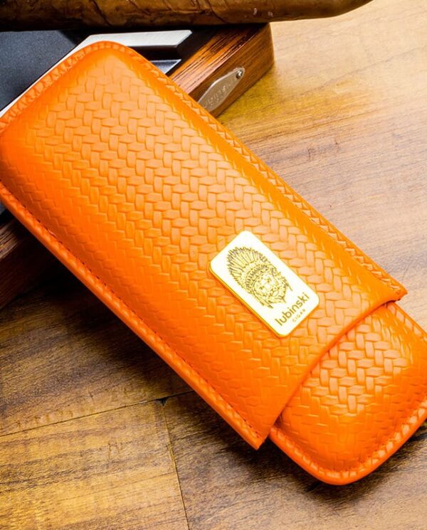 Orange woven leather case with a gold badge on the front, resting on a wood surface. It appears to be a compact accessory or sheath with stitched edges.