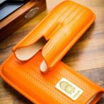 Orange woven leather glasses case, open to reveal beige interior on a wooden table, with a brand plaque visible in the corner.