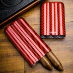 Red carbon-fiber cigar cases with pink stripes on a wooden surface; one opened to reveal two cigars, the other standing upright nearby.