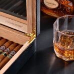 Open wooden cigar humidor with cigars inside, a gold hinge, and a glass of whiskey on a dark surface nearby, bottle labeled Hessenny in the background.