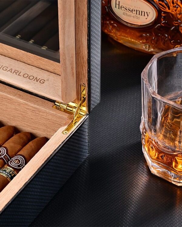 CH025-02 Open wooden cigar humidor with cigars inside, a gold hinge, and a glass of whiskey on a dark surface nearby, bottle labeled Hessenny in the background.