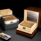 Luxury wooden cigar humidor with glass lid, open to reveal neatly packed cigars and a digital humidity gauge reading around 70%.