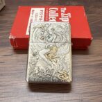 Engraved metal lighter showing a mermaid among sea creatures, placed on a wooden surface with a red box in the background.