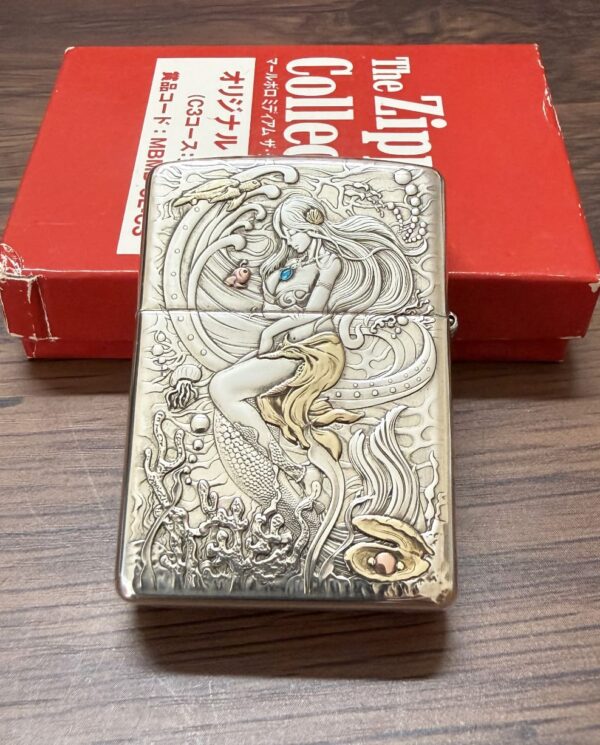 KZ003_01 Engraved metal lighter showing a mermaid among sea creatures, placed on a wooden surface with a red box in the background.