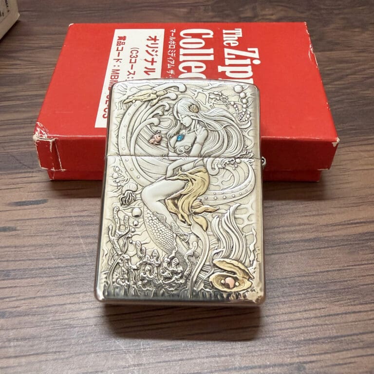 KZ003_01 Engraved metal lighter showing a mermaid among sea creatures, placed on a wooden surface with a red box in the background.