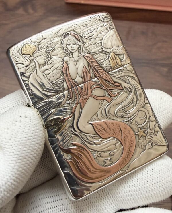 KZ003_08 Engraved metal lighter showing a mermaid with flowing hair and orange tail among seashells and waves.