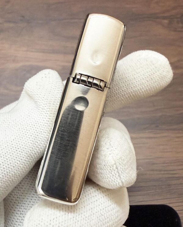 KZ003_14 Gloved hand holding a closed, silver metal lighter (Zippo-style) with a hinged lid, close-up view on a wooden surface.