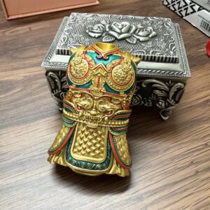 KZ364-01 Gold-toned ornate ceremonial mask/helmet figurine with red and teal accents and intricate raised patterns, resting on a wooden table beside a silver decorative box.