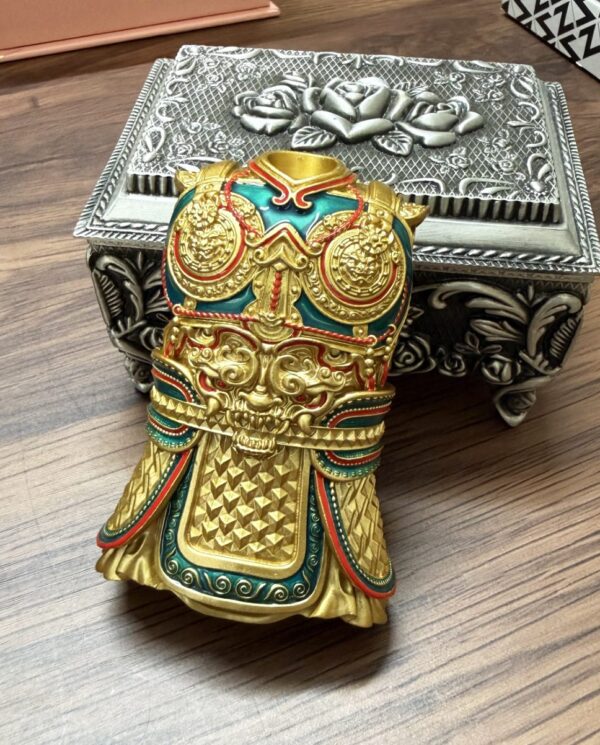 Gold-toned ornate ceremonial mask/helmet figurine with red and teal accents and intricate raised patterns, resting on a wooden table beside a silver decorative box.
