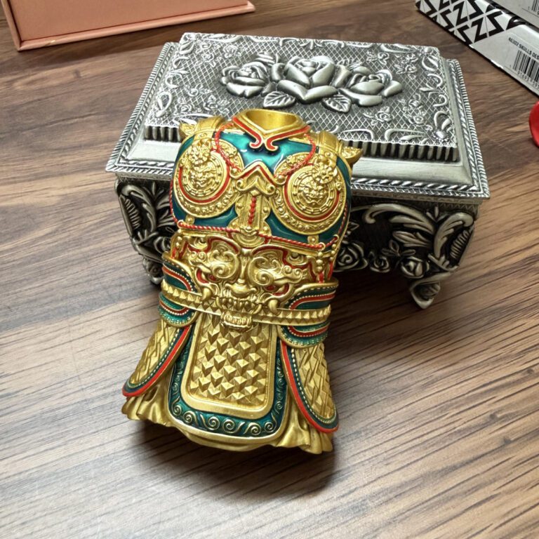 Gold-toned ornate ceremonial mask/helmet figurine with red and teal accents and intricate raised patterns, resting on a wooden table beside a silver decorative box.