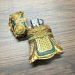 Ornate gold and teal metal lighter shaped like ceremonial armor, open to reveal the flint chamber inside a decorative grip area