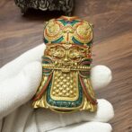Intricately designed gold-tone decorative mask or talisman with red and green enamel, held by a gloved hand, on a wooden surface.