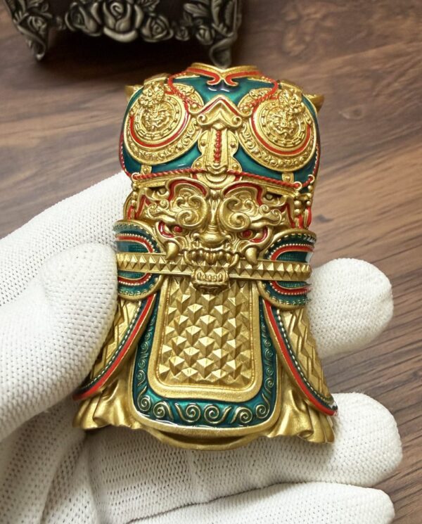 Intricately designed gold-tone decorative mask or talisman with red and green enamel, held by a gloved hand, on a wooden surface.