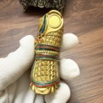 Close-up of a small, highly decorated gold figurine with scale-like engravings, turquoise and red enamel trim, being held by a gloved hand over a wooden surface.
