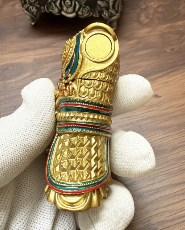 Close-up of a small, highly decorated gold figurine with scale-like engravings, turquoise and red enamel trim, being held by a gloved hand over a wooden surface.