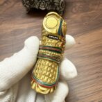 Gold decorative amulet in the shape of a bird with turquoise and red enamel bands, held by a gloved hand.