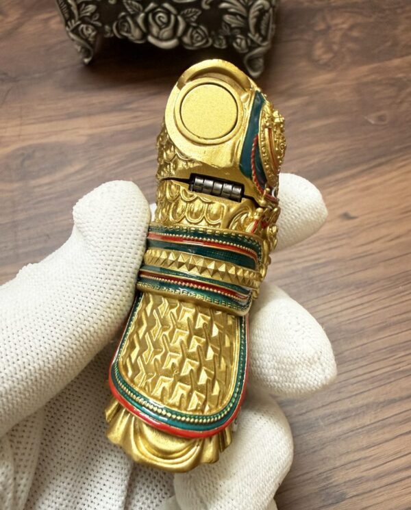 Gold decorative amulet in the shape of a bird with turquoise and red enamel bands, held by a gloved hand.