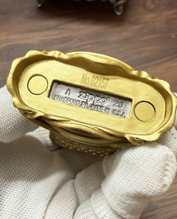 Gold-tone Zippo lighter bottom with serial No. 02157, Bradford, PA, Made in USA, held by a gloved hand.