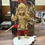 Small collectible figure of a warrior in ornate gold armor with red pants, holding a gold staff, standing on a white decorative base.