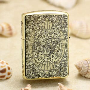 Gold Zippo lighter with intricate skull and rose engraving resting on sandy surface with seashells.