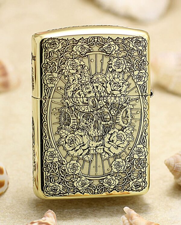 KZ365-01 Gold Zippo lighter with intricate skull and rose engraving resting on sandy surface with seashells.