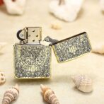 Gold engraved Zippo-style lighter opened, resting on a sandy beach with seashells nearby; vintage floral pattern on the case.
