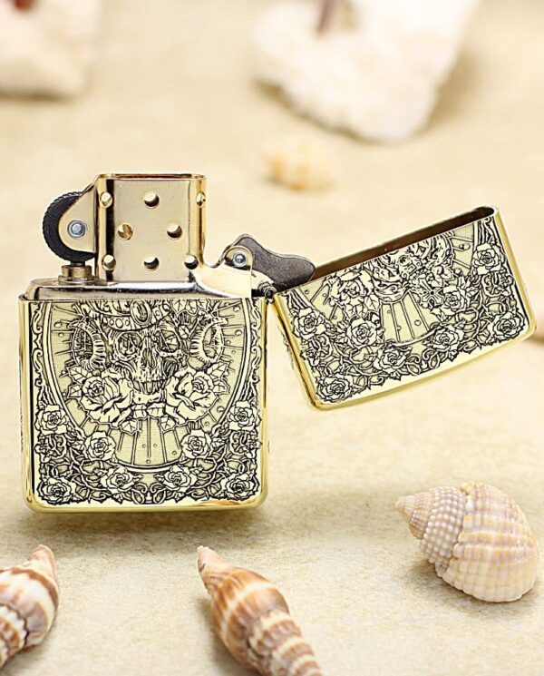 KZ365-03 Gold engraved Zippo-style lighter opened, resting on a sandy beach with seashells nearby; vintage floral pattern on the case.