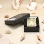 Gold engraved lighter in an open black box on a sandy beach with seashells nearby (presentation or product shot). -or- A gold-engraved lighter displayed in its black box on a beach-themed surface.